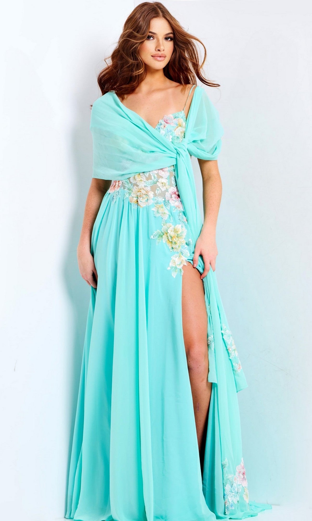A woman in the Jovani 49633 long prom dress—flowing aqua blue with floral embroidery, appliques, a thigh-high slit, sheer corset bodice, and matching shawl—stands against a plain light background.